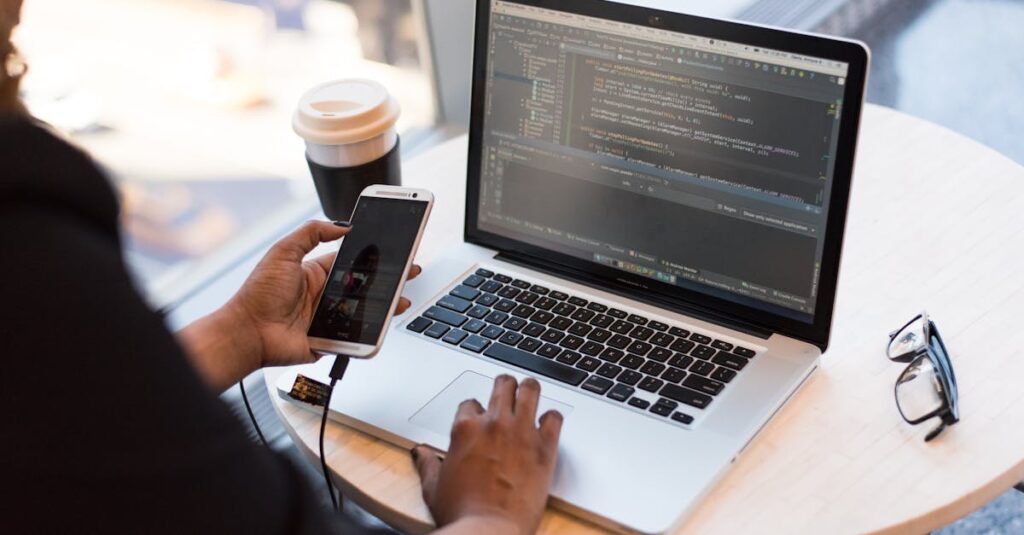 Developer working remotely, coding on a laptop with phone in hand, showcasing modern work culture.